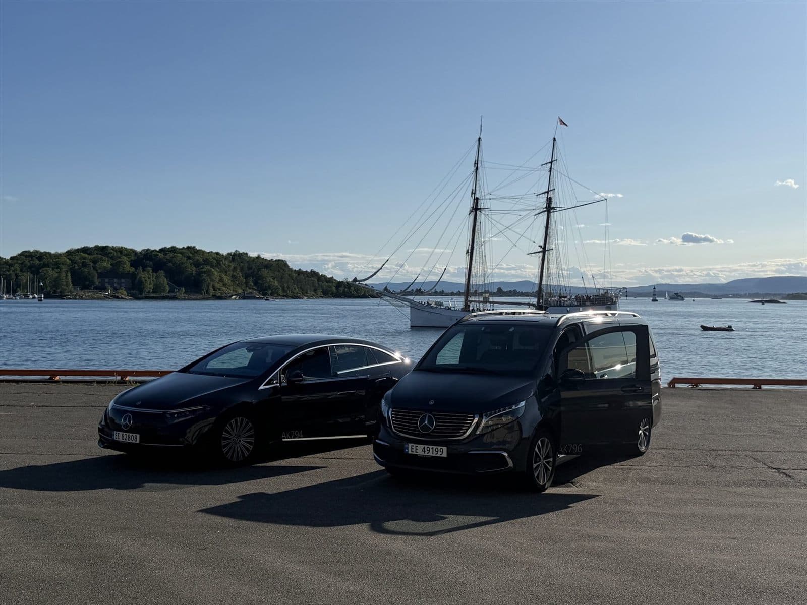 En svart Mercedes EQS og en Mercedes EQV parkert på en kai ved Oslofjorden, med et seilskip i bakgrunnen.