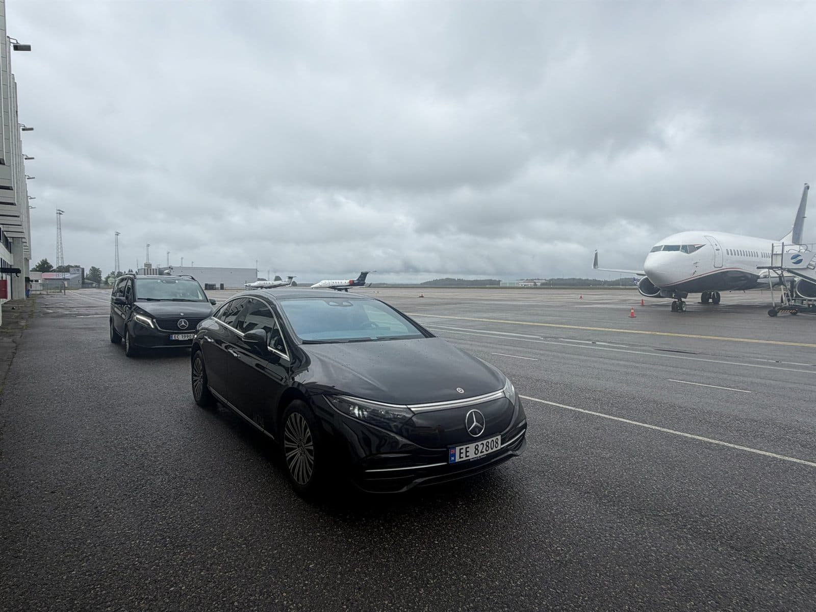 En svart Mercedes EQS foran en Mercedes EQV på en flyplass, med fly i bakgrunnen.